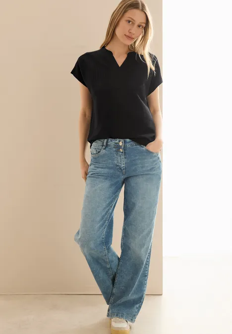 Frau trägt schwarzes Shirt und weite Jeans vor hellem Hintergrund.
